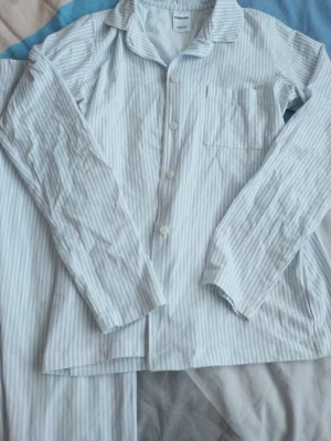 Randig pyjamas från KappAhl - Klassisk tvådelad pyjamas från KappAhl i ljusblått och vitt med tunna vertikala ränder. Skjortan har lång ärm, bröstficka och knappar framtill. Byxorna är raka och lösa. Tillverkad i mjukt bomullstyg för extra komfort.