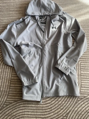 Ljusgrå vindjacka från Under Armour - Snygg ljusgrå vindjacka från Under Armour med huva och dragkedja framtill. Jackan har elastiska muddar vid ärmsluten, två sidofickor och logga på bröstet. Tillverkad i lätt och vindtätt material, perfekt för blåsiga dagar. PRIS KAN DISKUTERAS 