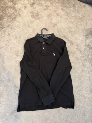 Skjorta från Polo Ralph Lauren - Klassisk svart långärmad skjorta från Polo Ralph Lauren med vit broderad logga på bröstet. Skjortan har krage, knappar framtill och är tillverkad i mjuk bomull. Perfekt för en clean och stilren look. Den sitter lite mer som M men passar båda