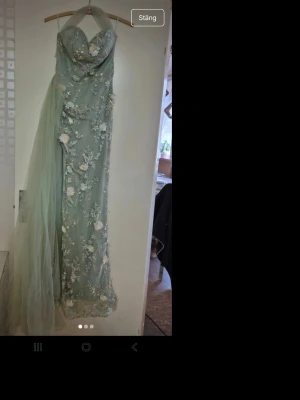 Grön långklänning med blommor - Säljer en elegant grön långklänning med blommigt mönster och broderade detaljer. Klänningen är axelbandslös och har en vacker tyllsläp på sidan som ger extra wow-känsla. Perfekt för dig som vill sticka ut på bal eller fest!