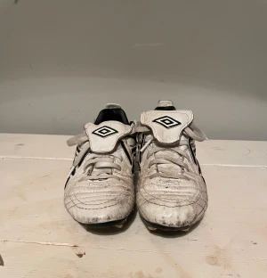 Vita Umbro fotbollsskor med dobbar - Säljer ett par klassiska vita fotbollsskor från Umbro med svarta detaljer och logga på sidan och plösen. Skorna har snörning, dobbar för gräsplan och quiltad ovandel i syntetmaterial. 
