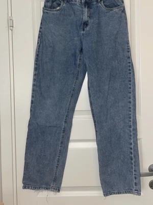 Blå raka jeans med rå kant - Ett par klassiska blå jeans med rak passform och hög midja. Jeansen har fem fickor och rå, ofållad kant nedtill för en trendig look. Tillverkade i kraftigt denimtyg som ger en autentisk vintagekänsla.