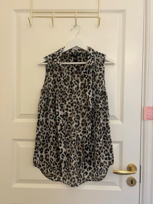 Ärmlös leopardmönstrad blus H&M - Snygg ärmlös blus från H&M med leopardmönster i beige, brunt och svart. Blusen har krage och knäppning framtill, samt en lös och luftig passform. Perfekt för dig som vill sticka ut med ett djurmönster.