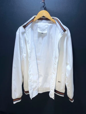 Vit vindjacka med randiga muddar - Snygg vit vindjacka med dragkedja och ribbade muddar i vitt, brunt och svart. Jackan har ståkrage och två sidofickor med dragkedja. Perfekt för en sportig och avslappnad stil. Tillverkad i lätt och vindtätt material.