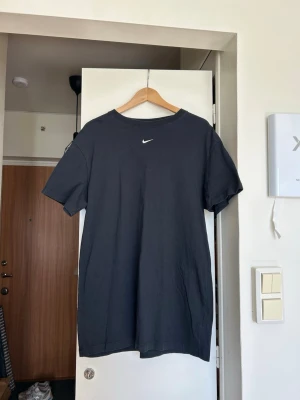 Svart Nike t-shirtklänning i bomull - En svart t-shirtklänning från Nike med klassisk rund hals och korta ärmar. Klänningen har en liten vit Nike-logga på bröstet och är gjord i mjuk bomull för en clean och enkel stil. Storlek M men skulle säga att det snarare är en S om man vill ha den lite baggy. 