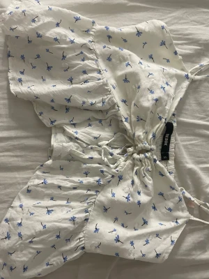 Vit blommig topp från Bik Bok - Supersöt vit topp från Bik Bok med små blå blommor, korta ärmar och snörning framtill. Toppen har en lätt croppad passform och tunna axelband. Perfekt för varma dagar och ger en fräsch sommarkänsla.
