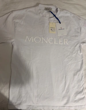 Vit Moncler t-shirt med logga - Säljer en vit t-shirt från Moncler med stor broderad logga på bröstet och en ficka med knapp och Moncler-märke på ärmen. Klassisk rund hals och korta ärmar. Tillverkad i mjuk bomull för en clean och exklusiv look.