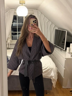 Svart quiltad kimono med bälte - Snygg svart quiltad kimono-inspirerad tröja med trekvartsärmar och knytband i midjan. Perfekt att slänga över en topp för en chill men ändå stilren look. Materialet är mjukt och ger en avslappnad vibe.
