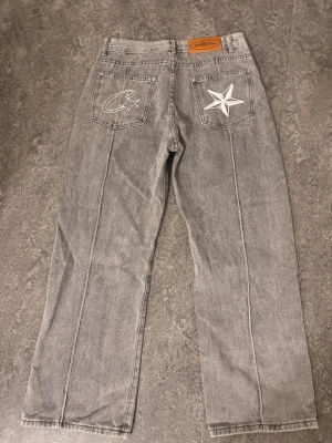  Grå Corteiz jeans - Säljer ett par grå raka jeans från Corteiz med unika broderade detaljer på bakfickorna – en stjärna och ett C. Jeansen har hög midja, klassisk femficksdesign och kontrastsömmar. Materialet är slitstark denim i 100% bomull.