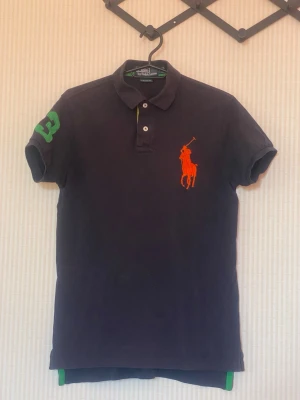 Svart pikétröja från Ralph Lauren - Klassisk svart pikétröja från Ralph Lauren med orange logga på bröstet och grönt nummer på ärmen. Tröjan har krage, knappar framtill och korta ärmar. Perfekt för en sportig och stilren look.