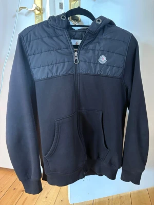 Svart hoodie från Moncler med dragkedja - Snygg svart hoodie från Moncler med quiltade detaljer på axlar och huva. Dragkedja framtill, fickor och ribbade muddar. Ikonisk Moncler-logga på bröstet. Perfekt för dig som vill ha en stilren och exklusiv look. Kan fraktas eller mötas upp i närheten av Skövde mvh Lukas.