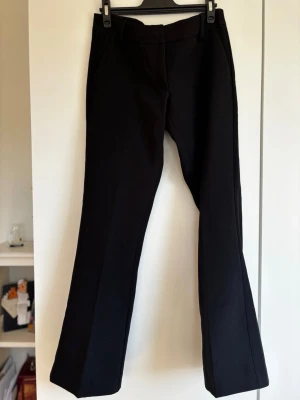 Svarta kostymbyxor med utsvängda ben - Snygga svarta kostymbyxor med utsvängda ben och pressveck. Byxorna har hög midja och diskreta fickor fram samt passpoalfickor bak. Perfekta för en clean och stilren look. Materialet ser ut att vara en mjuk och stretchig mix, vilket ger skön passform. I petit kollektionen. Ej använd. 