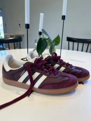 Vinröda Adidas Samba sneakers - Snygga Adidas Samba sneakers i vinrött läder med klassiska vita ränder på sidorna och gummisula. Skorna har vinröda skosnören och vit hälkappa. Perfekta för dig som vill ha en ikonisk och stilren sneaker med retrovibbar.