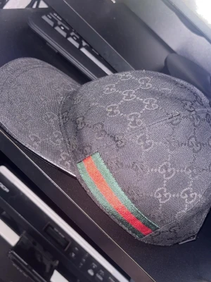 Svart keps från Gucci med GG-mönster - Säljer en svart keps från Gucci med det klassiska GG-monogrammönstret över hela kepsen. Kepsen har böjd skärm, justerbart spänne bak och diskreta röda och gröna detaljer på sidorna. Tillverkad i canvasmaterial med exklusiv känsla.