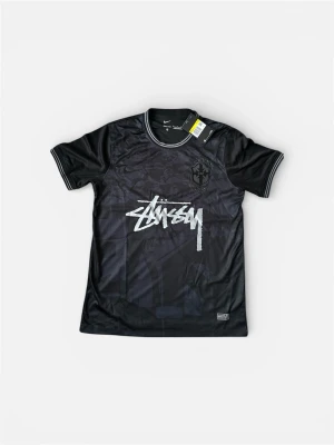 Svart Stüssy x Nike t-shirt ☀️ - Snygg svart t-shirt från Stüssy x Nike med korta ärmar och vit Stüssy-logga framtill. T-shirten har diskreta mönsterdetaljer och broderad patch på bröstet. Tillverkad i mjuk polyester som andas, perfekt för en streetwear-look.