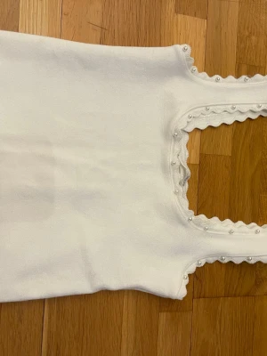 Vit linnetopp med pärlor - Supersöt vit linnetopp med dekorativa pärlor längs den vågiga kanten vid halsringningen och axelbanden. Perfekt att styla till jeans eller kjol för en fräsch look.