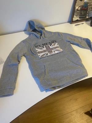 Grå hoodie med brittisk flagga - Säljer en grå hoodie med tryck av brittiska flaggan och texten 'MY HEART BEATS TO A BRITISH TUNE' på bröstet. Hoodien har huva med dragsko och en klassisk känguruficka framtill. Perfekt för dig som gillar streetwear och vill sticka ut lite extra. Storlek L men skulle förmodligen också passa M. Aldrig använd, helt ny. 