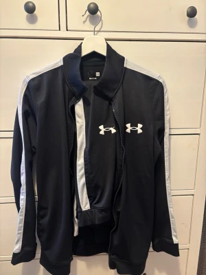 Svart träningset från Under Armour - Snygg svart träningset från Under Armour med vita ränder längs ärmarna och logga på bröstet. Jackan har dragkedja framtill, hög krage och muddar vid ärmslut och midja. Perfekt för gymmet eller löprundan.