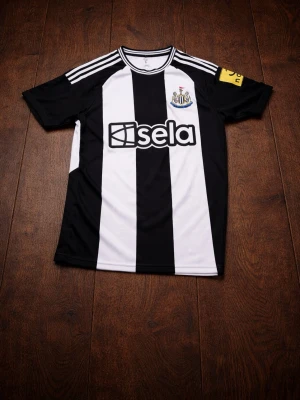 Newcastle United matchtröja svart/vit - Snygg Newcastle United matchtröja från Adidas i svart och vitt med klassiska ränder. Tröjan har klubbmärke på bröstet, sponsorlogga 'sela' och gula detaljer på ärmen. På ryggen står det ISAK och nummer 14 i rött. Perfekt för fotbollsfans!