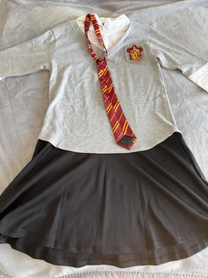 Gryffindor skoluniform cosplaydräkt - Cosplaydräkt inspirerad av Gryffindor från Harry Potter. Setet består av en grå tröja med fastsydd vit skjorta, svart kjol och en röd/gul slips med Gryffindor-emblem på bröstet. Perfekt för maskerad eller cosplayevent!