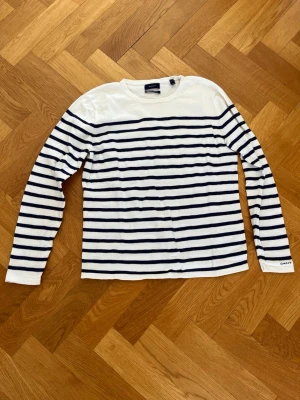 Randig långärmad tröja från GANT - Säljer en klassisk långärmad tröja från GANT i vitt med mörkblå horisontella ränder. Tröjan har rund halsringning och en relaxed passform. Tillverkad i mjukt bomullsmaterial och har en liten GANT-logga på ärmslutet.