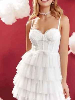 Studentklänning - Supersöt vit studentklänning med tunna axelband, spetsdetaljer upptill och flera lager volanger i kjolen. Klänningen har en feminin och romantisk look, perfekt för sommarens alla tillfällen. Materialet känns lätt och luftigt, har aldrig använts 