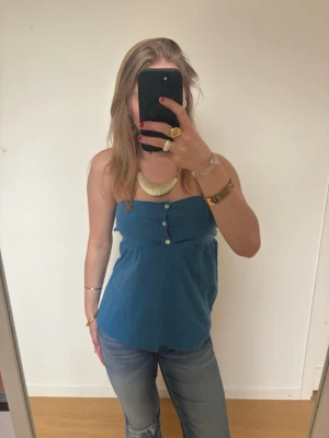 Blå axelbandslös topp med knappar - Säljer en blå axelbandslös topp med tre dekorativa knappar framtill. Toppen, är sydd av mig, och säljs billigt på grund av att jag inte blev nöjd med sömmarna ex se sista bild. Kolla gärna på mina andra annonser. Ställ gärna frågor!🩷