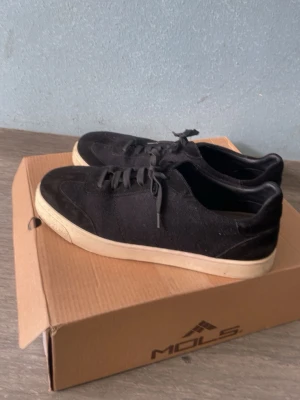 Svarta sneakers från MOLS - Stilrena svarta sneakers från MOLS med lågt skaft och vita sulor. Skorna har klassisk snörning och är tillverkade i tygmaterial som ger en avslappnad look. Perfekta för dig som gillar enkel och clean stil.