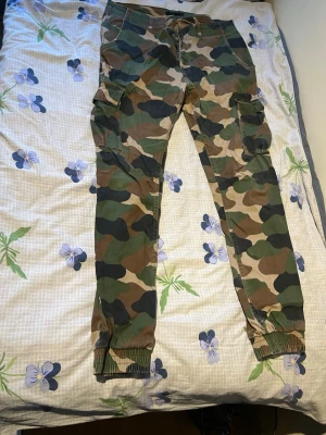 Cargobyxor i camomönster - Säljer ett par cargobyxor med klassiskt camouflagemönster i grönt, brunt och beige. Byxorna har flera praktiska fickor på sidorna och resår vid bensluten för en snygg passform. Perfekta för en streetwear-look eller till vardagsoutfiten.