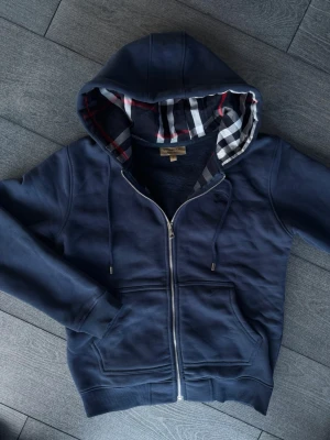 Marinblå hoodie från Burberry - Snygg marinblå hoodie från Burberry med dragkedja och klassiskt rutigt foder i huvan och längs insidan. Hoodien har två fickor framtill, ribbade muddar och justerbar dragsko i huvan. Perfekt för en chill och stilren look.