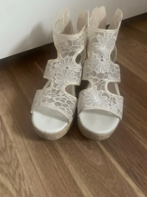 Vita kilklackade spets-sandaler - Säljer ett par vita sandaler från DinSko med kilklack och öppen tå. Ovandelen är i spets med blommönster och har flera utskärningar för en luftig känsla. Klacken är täckt med flätad jute för en somrig look. Perfekta för dig som vill ha något extra till outfiten.