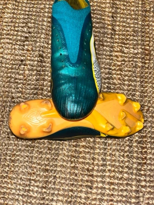 Puma fotbollsskor blå/gula - Snygga fotbollsskor från Puma i blått och gult med vita detaljer och grafiskt mönster på sidan. Skorna har låg profil, strumpliknande passform och fasta dobbar i plast för spel på gräs. Ovandelen är i syntetmaterial för bra bollkontroll.