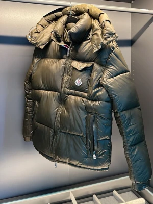 Olivgrön dunjacka från Moncler - Snygg olivgrön dunjacka från Moncler med stor huva och quiltad design. Jackan har dragkedja framtill, två sidofickor med dragkedja och en bröstficka med lock och Moncler-logga. Perfekt för kalla vinterdagar och riktigt trendig look.