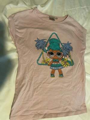 Ljusrosa LOL Surprise t-shirt barn st 122 - Söt ljusrosa t-shirt från LOL Surprise med färgglad tryck framtill som visar en tecknad figur, palmer och blommor. T-shirten har rund hals och korta ärmar, perfekt för dig som gillar lekfulla motiv och mjuka material. Barn storlek 122
