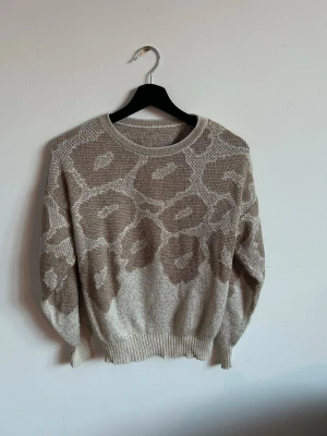 Beige stickad tröja med leopard mönster - Stickad tröja i beige och ljusbrunt med stort leopardmönster. Tröjan har rund halsringning, långa ärmar och ribbade muddar vid ärmslut och nederkant. Perfekt för dig som gillar en avslappnad och trendig stil. Glittrar och är lite guldig!