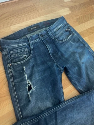 Replay anbass - Säljer ett par blå jeans från Replay med slitningar och hål på vänster lår. Jeansen har fem fickor, normal midja och raka ben. Klassisk tvättad denim-look med detaljerade sömmar och Replay-logga på bakfickan. Perfekta för en avslappnad streetstil. W29 L32