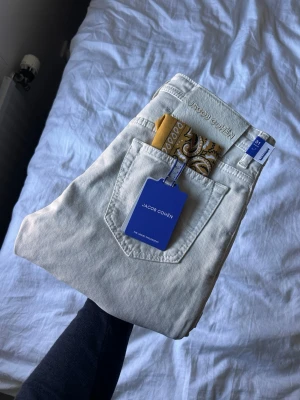 Beiga jeans från Jacob Cohën - Snygga beiga/krämvita jeans från Jacob Cohën med klassisk femficksmodell och diskret logga på bakfickan. Tillverkade i mjukt denimtyg och levereras med en gul paisleymönstrad näsduk i bakfickan. Perfekta för en clean och stilren look.