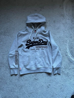 Superdry hoodie - Snygg grå hoodie från Superdry med stor logga och japanskt tryck på bröstet samt ärmen. Klassisk känguruficka framtill och justerbar huva med vita snören. Tillverkad i mjukt sweatshirtmaterial, perfekt för en chill och avslappnad stil. PRISET KAN DISKUTERAS VID SNABB AFFÄR.