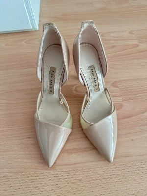 Beige lackpumps från Zara Basic - Snygga beige pumps från Zara Basic med spetsig tå och lackad finish. Skorna har öppen sida och låg klack, vilket ger en elegant och modern look. Tillverkade i syntetmaterial med en stilren design som passar till många outfits.
