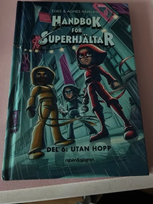 Handbok för Superhjältar Del 6: Utan hopp - Handbok för Superhjältar Del 6: Utan hopp av Elias & Agnes Våhlund. Boken har ett färgstarkt omslag med tecknade superhjältar i rött, gult och blått mot en stadsmiljö. Inbunden bok med spännande äventyr om Röda Masken och hennes vänner.