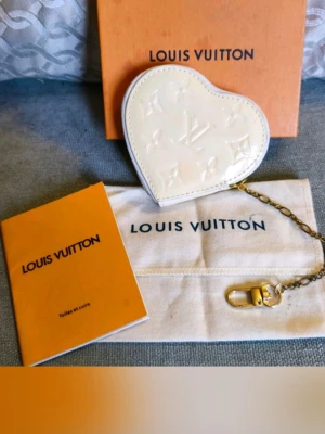 Vit hjärtformad coin purse  - Säljer en superfin hjärtformad coin purse från i Vernis med det klassiska monogramme.. Den har guldfärgad kedja och karbinhake, perfekt att fästa i väskan. Kommer med originalbox och dustbag. Har en missfärgning på baksidan där av lågt pris 