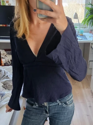 Mörkblå omlottblus med knytband / Tie top - Snygg mörkblå omlottblus med djup v-ringning och långa ärmar. Blusen har ett knytband i midjan som ger en fin siluett och är tillverkad i ett lätt, strukturerat tyg. Perfekt att styla med jeans eller kjol för en trendig look.