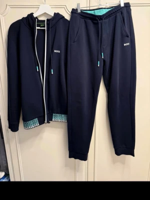 Mörkblå set från BOSS - Snyggt mörkblått set från BOSS med zip-hoodie och matchande byxor. Setet har turkosa detaljer och BOSS-logga på bröstet och bakfickan. Mjukt material, perfekt för chill dagar hemma. Fodret har ett mönster med BOSS-tryck i turkos och vitt.