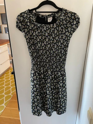 Blommig mörkblå kortklänning - Söt mörkblå kortklänning med små vita och gula blommor och fickor. Klänningen har korta ärmar, rund halsringning och smockad midja som ger en fin siluett. Den är i stretch material i midjan och är lite insydd i ryggen men inget som syns utifrån