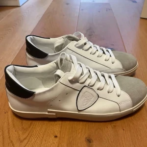 Philippe Model Vita Sneakers - Helt nya vita sneakers från Philippe Model med grå mockadetalj på tån och svart hälparti. Skorna har klassisk låg profil, vita skosnören och en sköldlogga på sidan. Tillverkade i läder och mocka för en clean och stilren look.