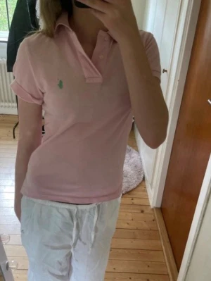 Rosa pikétröja från Ralph Lauren - Säljer en klassisk rosa pikétröja från Ralph Lauren med den ikoniska gröna loggan broderad på bröstet. Tröjan har korta ärmar, krage och knappar framtill. Perfekt för en avslappnad men ändå stilren look. Tror det är i storlek 152  men kan ha fel 
