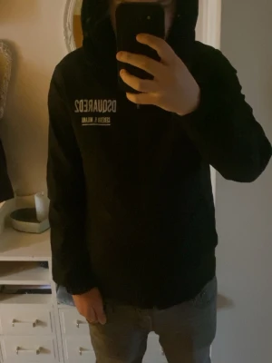 Svart hoodie från Dsquared2 - Snygg svart hoodie från Dsquared2 med vit logga och text både fram och bak. Klassisk huva och raka ärmar. Perfekt för dig som gillar streetwear och vill ha en stilren look. Materialet ser ut att vara mjukt och bekvämt, troligen bomull eller bomullsblandning.