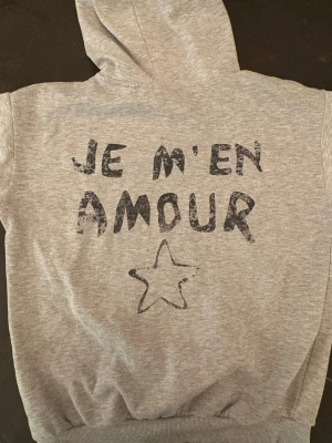 Grå hoodie med tryck 'JE M'EN AMOUR' - Jag säljer en jättefin ljusgrå hoddie med trycket JE M'EN AMOUR, den är ifrån Gina tricot och har använts 1-2 gånger💕hoddien är storlek 158/164