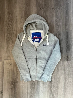 Grå zip hoodie från Superdry - Snygg grå hoodie från Superdry Orange Label med dragkedja, huva och vita dragsnören🤗 Passar nån som har stor Xs eller S(tecken på användning syns på bilderna). Hör av er vid funderingar samt snabbaffär🙌🏻