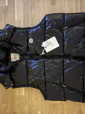 Svart dunväst från Moncler - Snygg svart dunväst från Moncler med glansig finish och avtagbar huva. Västen har två dragkedjeförsedda fickor och klassisk Moncler-logga på bröstet. Perfekt för lager-på-lager och streetwear-vibe. Materialet är dunfyllning och yttertyget är blankt.